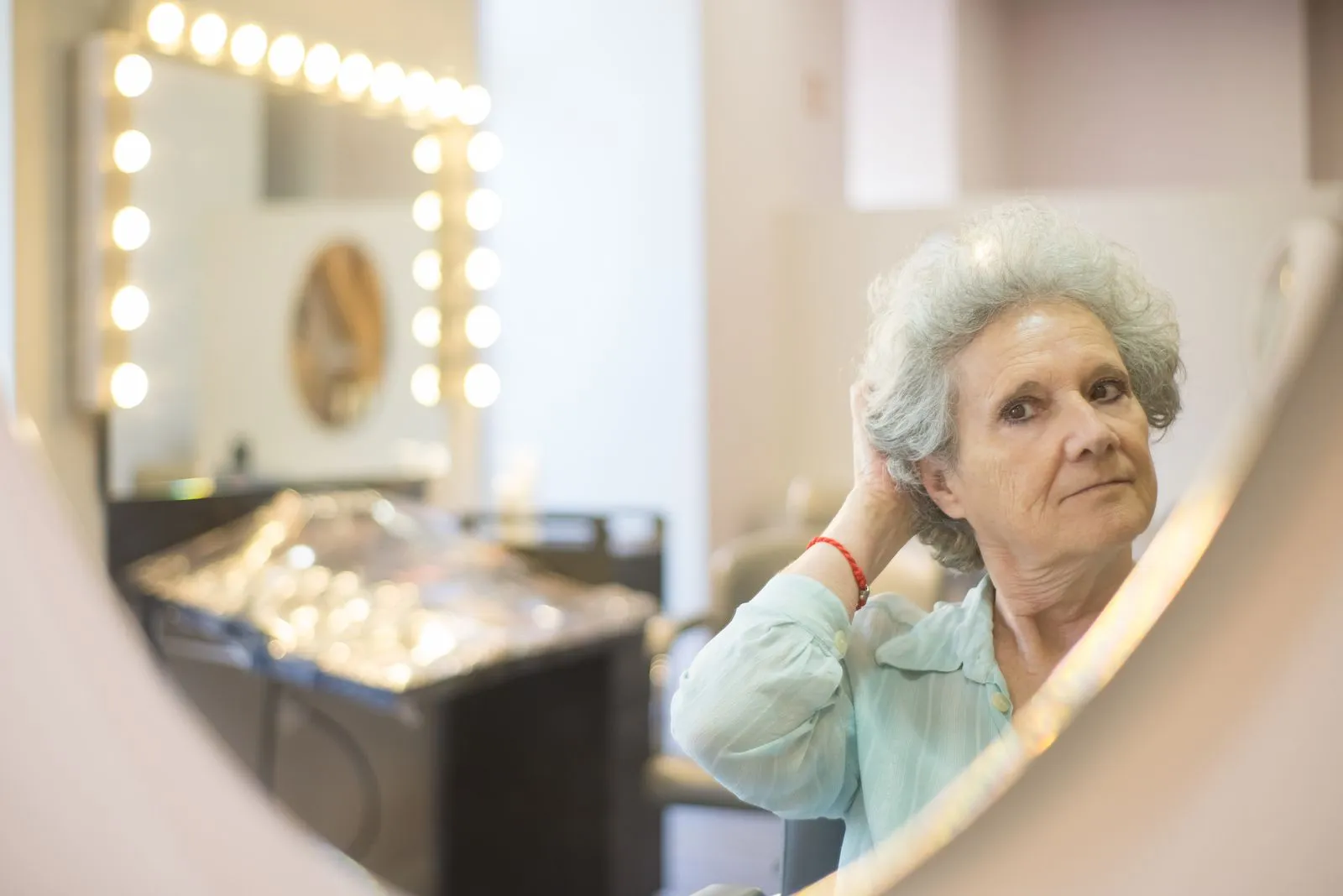 Senior woman adjusting her hair while looking into a mirror at a well-lit salon.